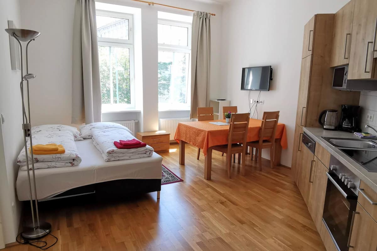 A bedroom with a bed, bedside lamp, and a window with curtains. There is a small table with chairs and a television mounted on the wall.