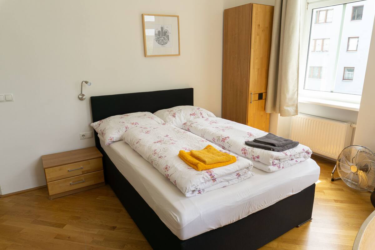 A neatly made bed with floral bedding, a small wooden nightstand, a framed picture on the wall, and a window with curtains.