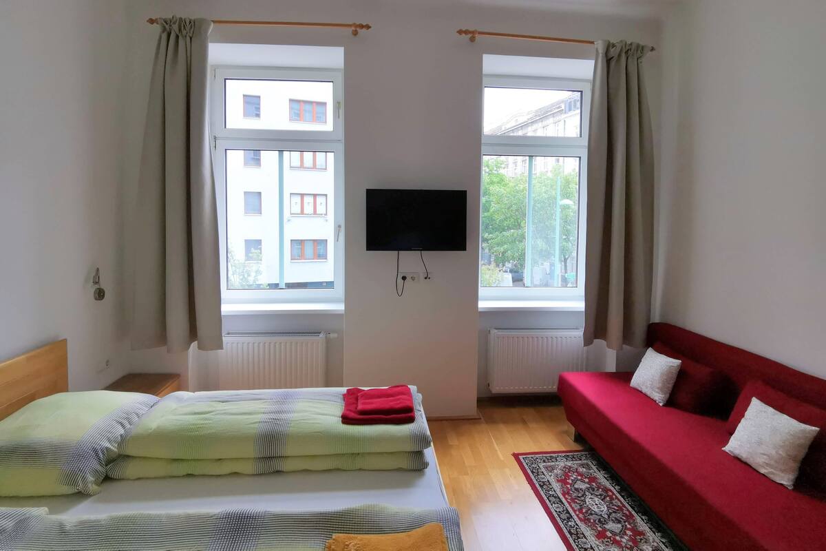 A room with a bed, a red sofa, a TV, and two windows with curtains.