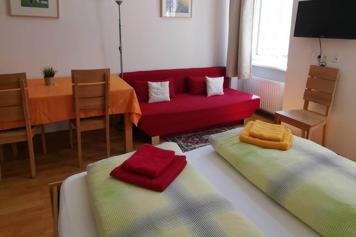 A room with a bed, a red sofa, a dining table with chairs, and a TV on the wall.