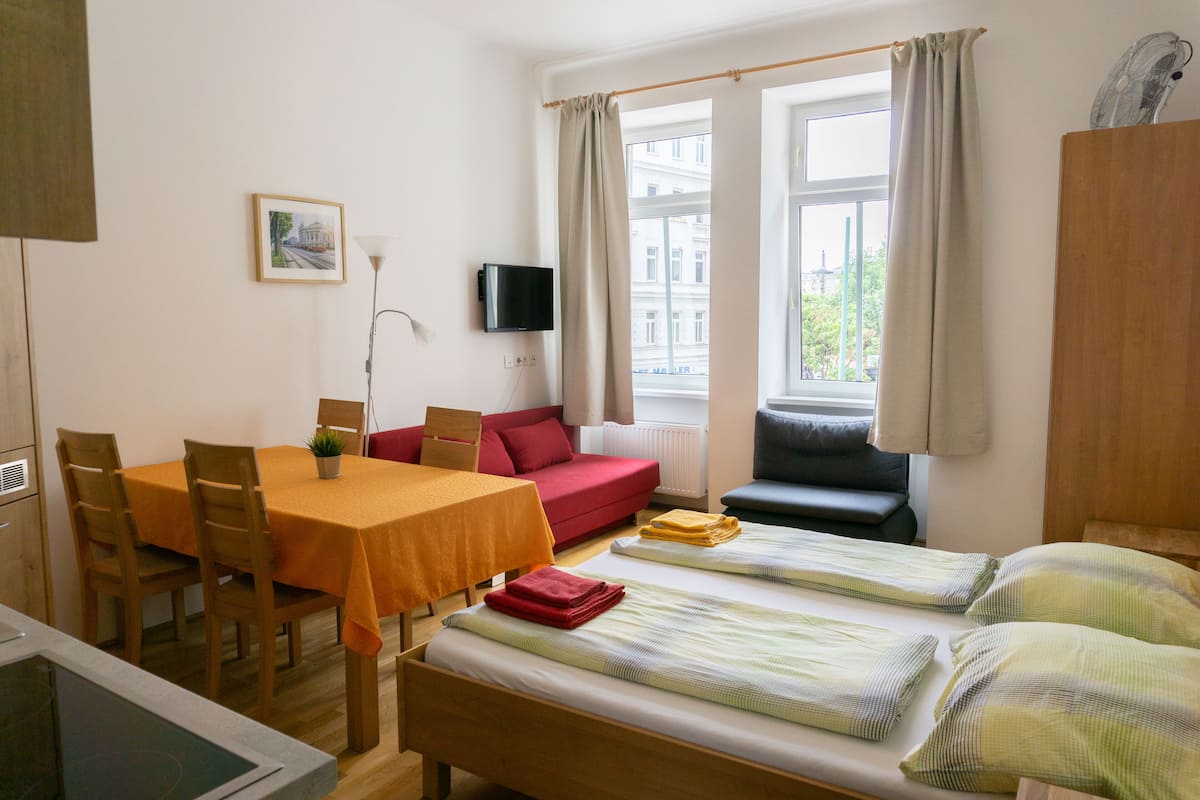 A room with a bed, a sofa, a table with chairs, a TV, and a window with curtains.