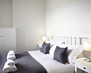 Un lit bien fait, avec du linge de lit blanc et des oreillers gris, une table de chevet avec une lampe et une armoire blanche.
