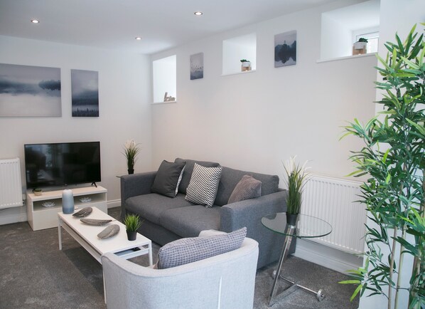 Un salon moderne avec un canapé gris, un téléviseur à écran plat, une table basse en verre et une petite table d’appoint blanche agrémentée d’une plante.