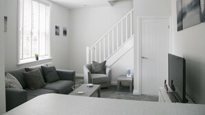 Un salon moderne avec un canapé gris, une table basse et un téléviseur. Un escalier mène à un autre niveau, et une porte se trouve sur la droite.