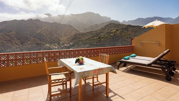 A balcony with a table, chairs, and a sun lounger overlooking a mountainous landscape.