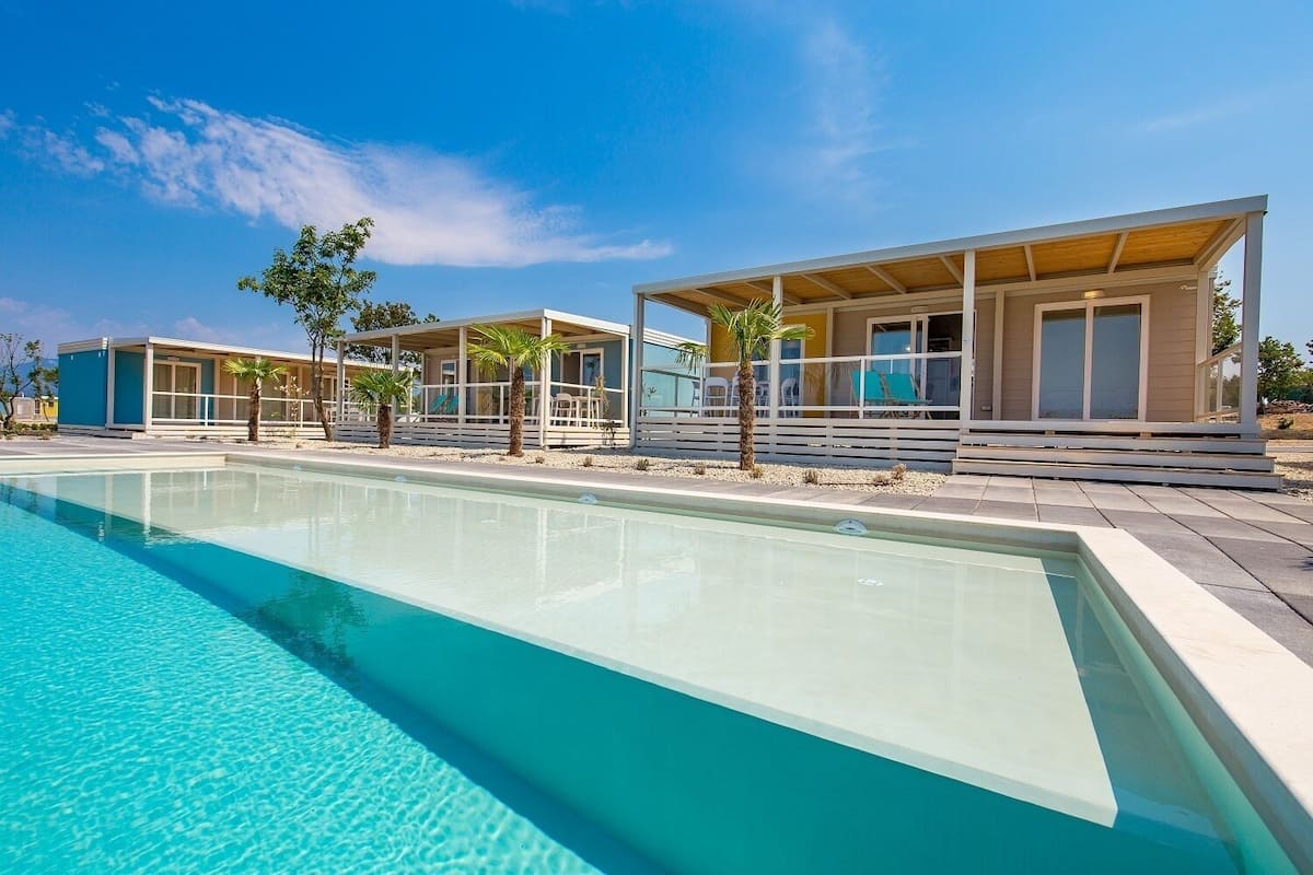 Un espace moderne aménagé au bord d’une piscine à eau claire, jouxtant un bâtiment doté d’un patio couvert et d’un espace salon.