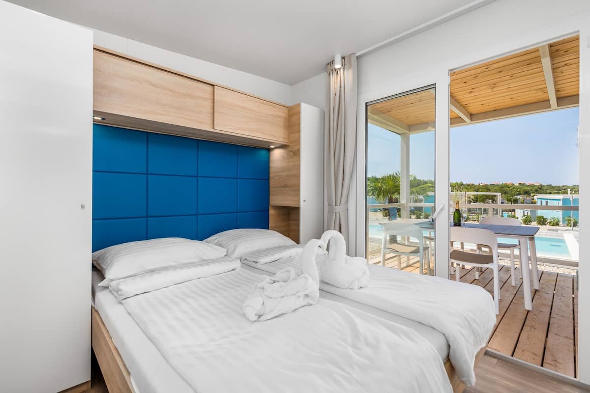 Une chambre d’hôtel moderne dotée d’un grand lit, d’une tête de lit en bois et offrant une vue sur une piscine et un espace de détente extérieur.