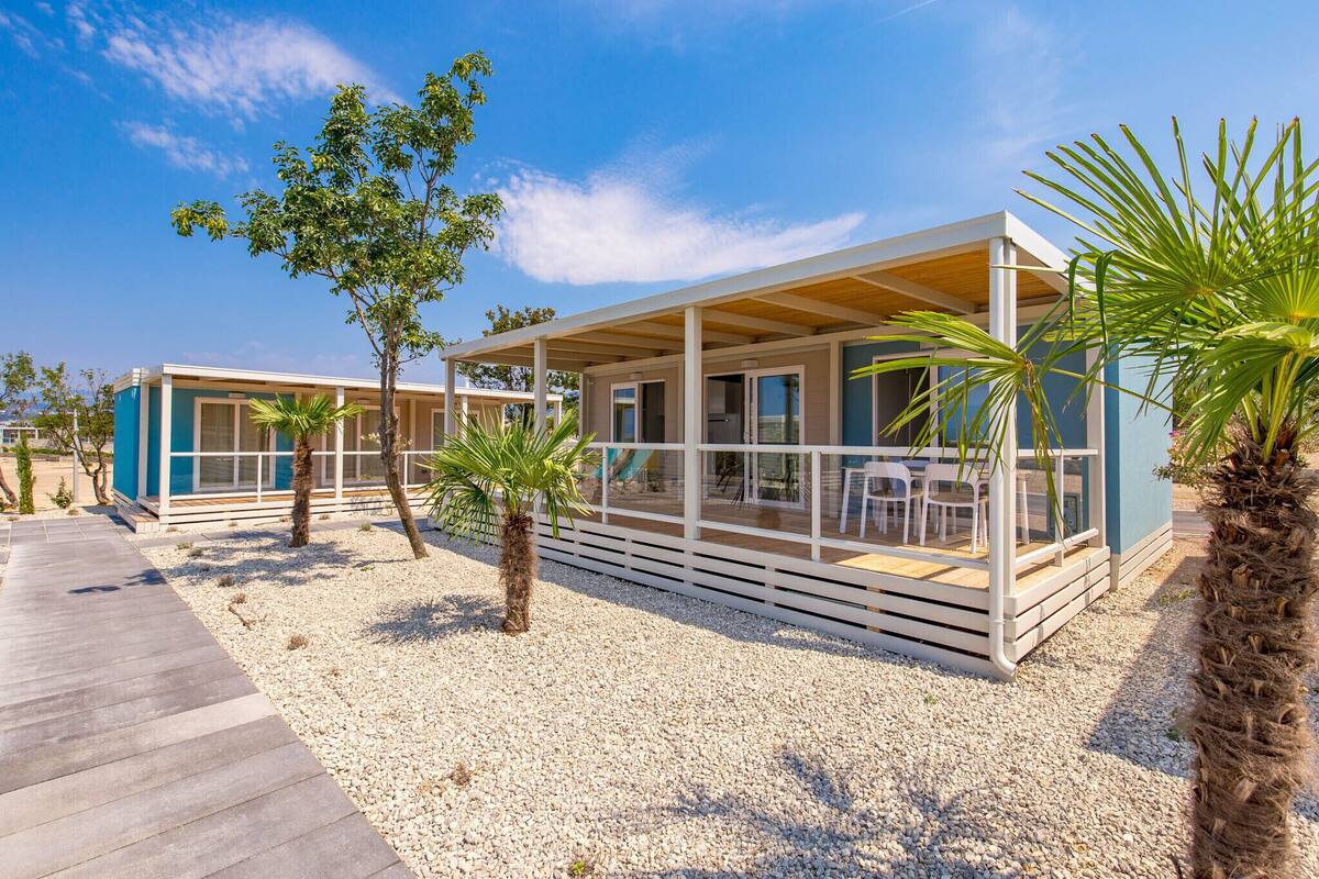 Un complexe hôtelier moderne situé en bord de mer, doté d’un espace extérieur couvert avec des sièges, de palmiers et d’un sentier en gravier.