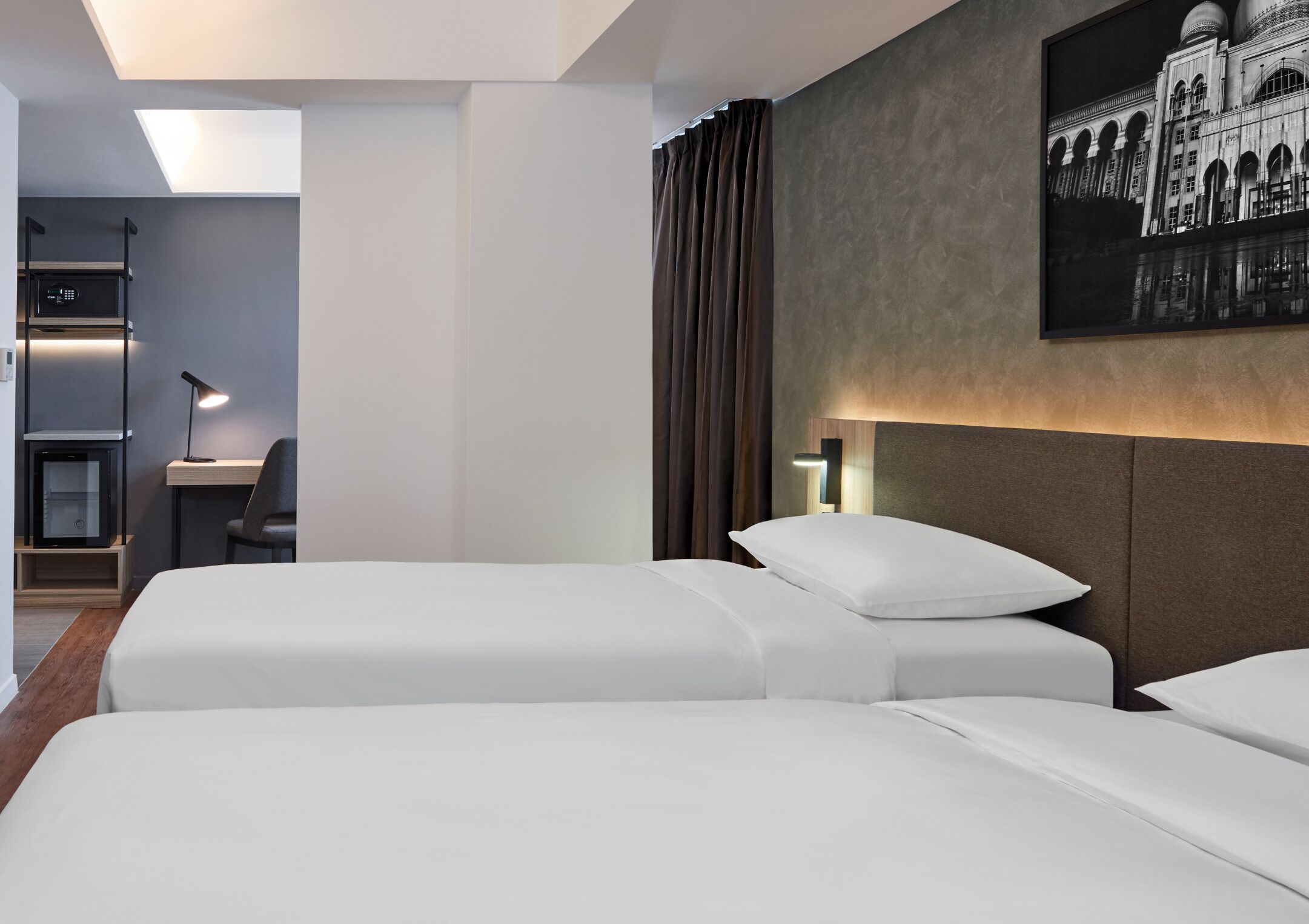 A hotel room with two beds, a desk, a chair, a lamp, and a wall-mounted picture.