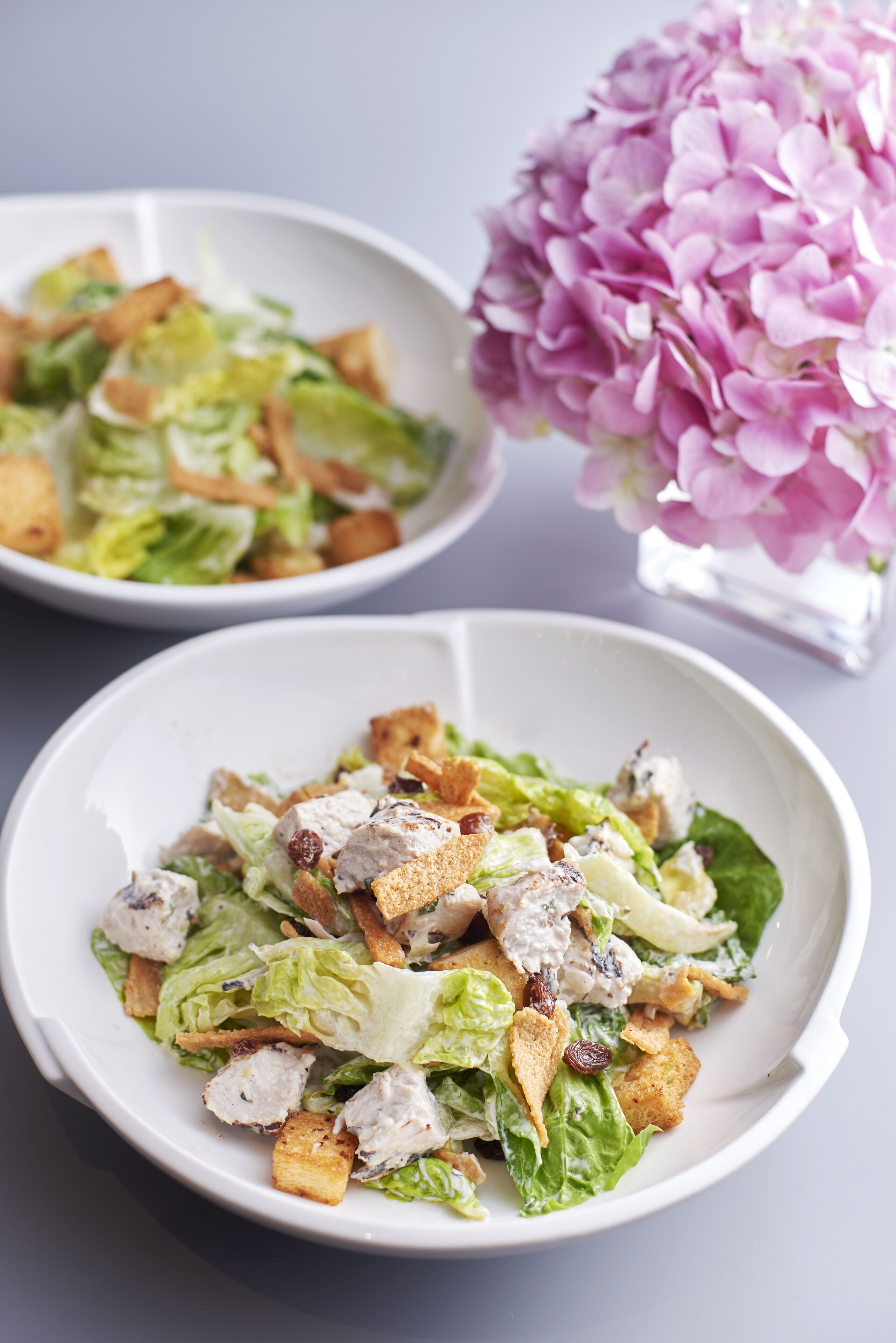 Deux salades césar, servies dans des bols blancs, garnies de croûtons et de laitue, sont posées sur une table. Un vase rempli de fleurs roses se trouve à l’arrière-plan.