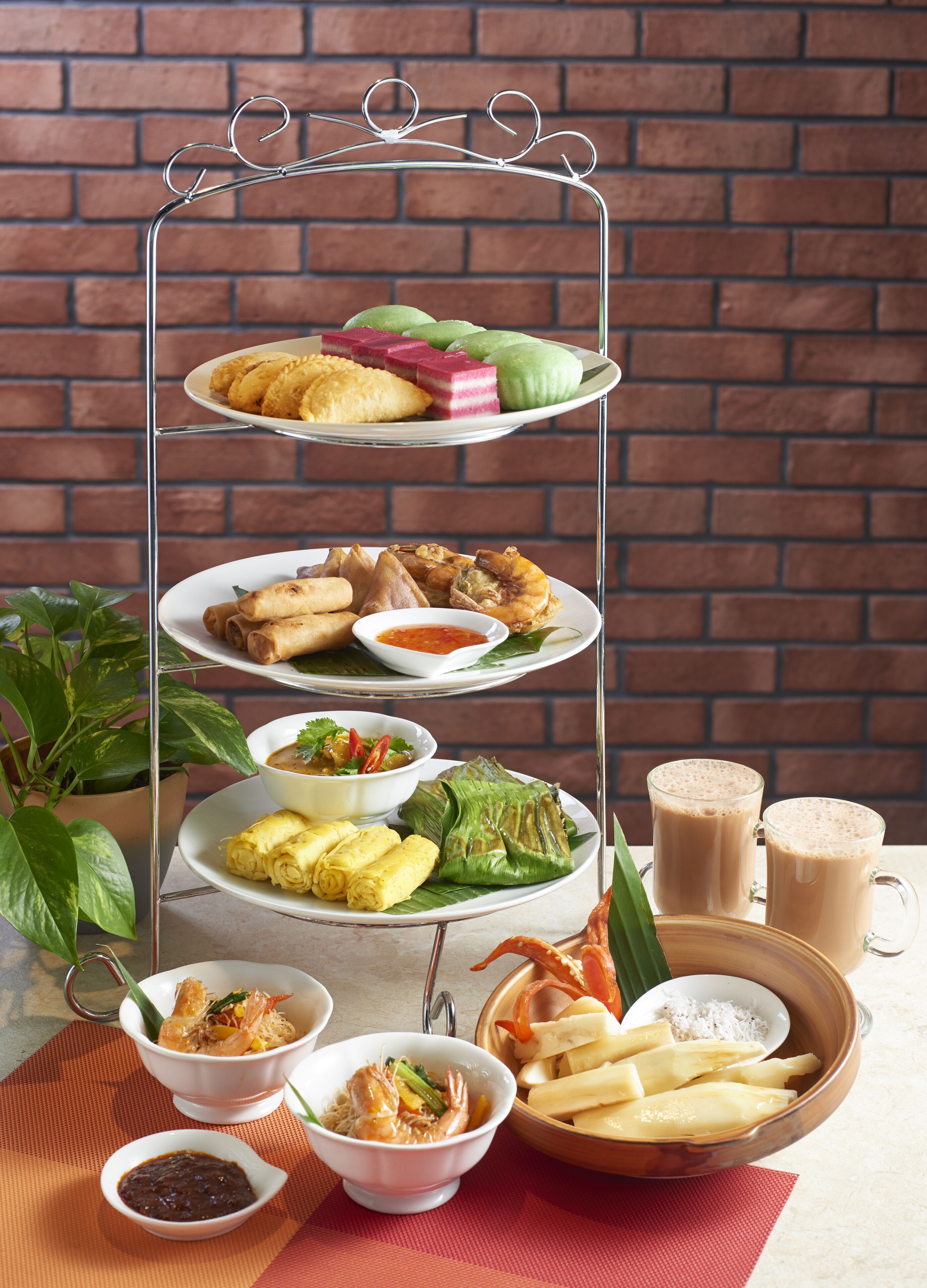 A tiered serving tray with various dishes, including fried items, a bowl of soup, and a plate of wrapped food, set against a brick wall background.