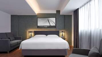 A modern hotel room with a large bed, a sofa, a TV mounted on the wall, and a window with curtains.
