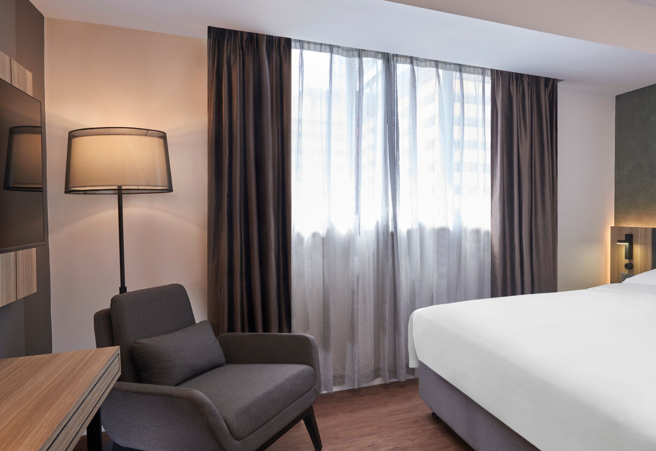 A hotel room with a bed, a chair, a desk, a lamp, and a window with curtains.