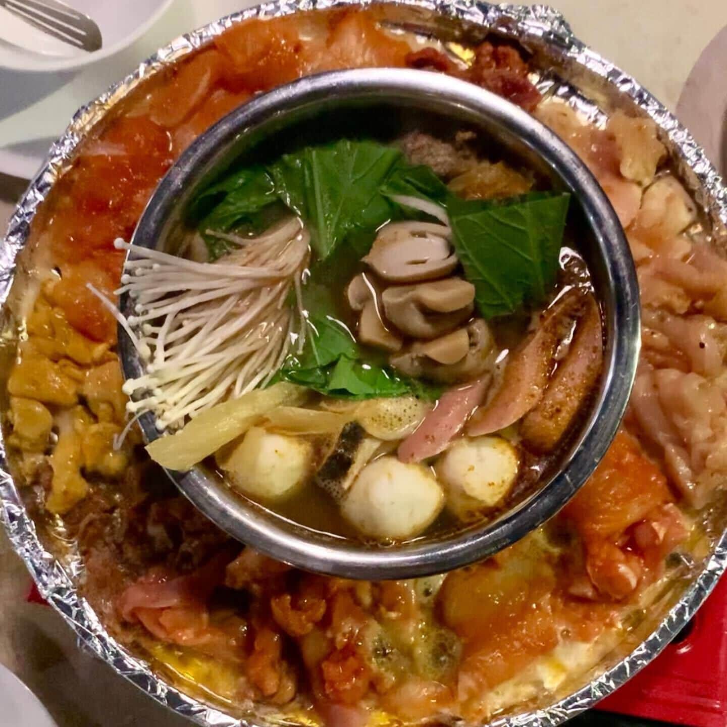 Un tazón de sopa con diversos ingredientes como hongos, tofu y verduras de hoja, servido en un tazón de metal sobre una bandeja cubierta con papel de aluminio.