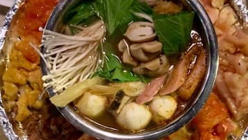 A bowl of soup with various ingredients like mushrooms, tofu, and leafy greens, served in a metal bowl on top of a foil-lined tray.