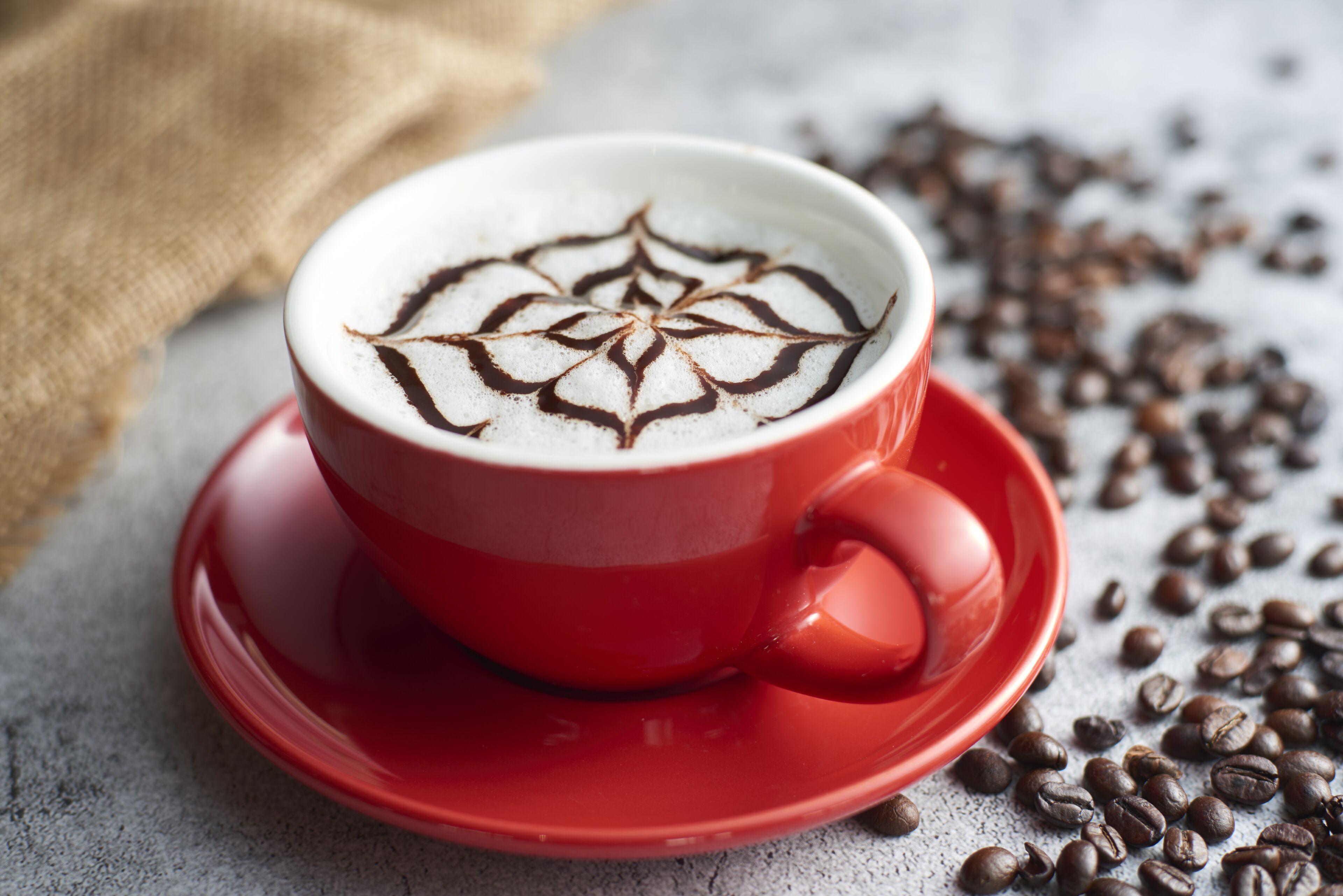 A red cup of coffee with latte art on a saucer, surrounded by coffee beans.
