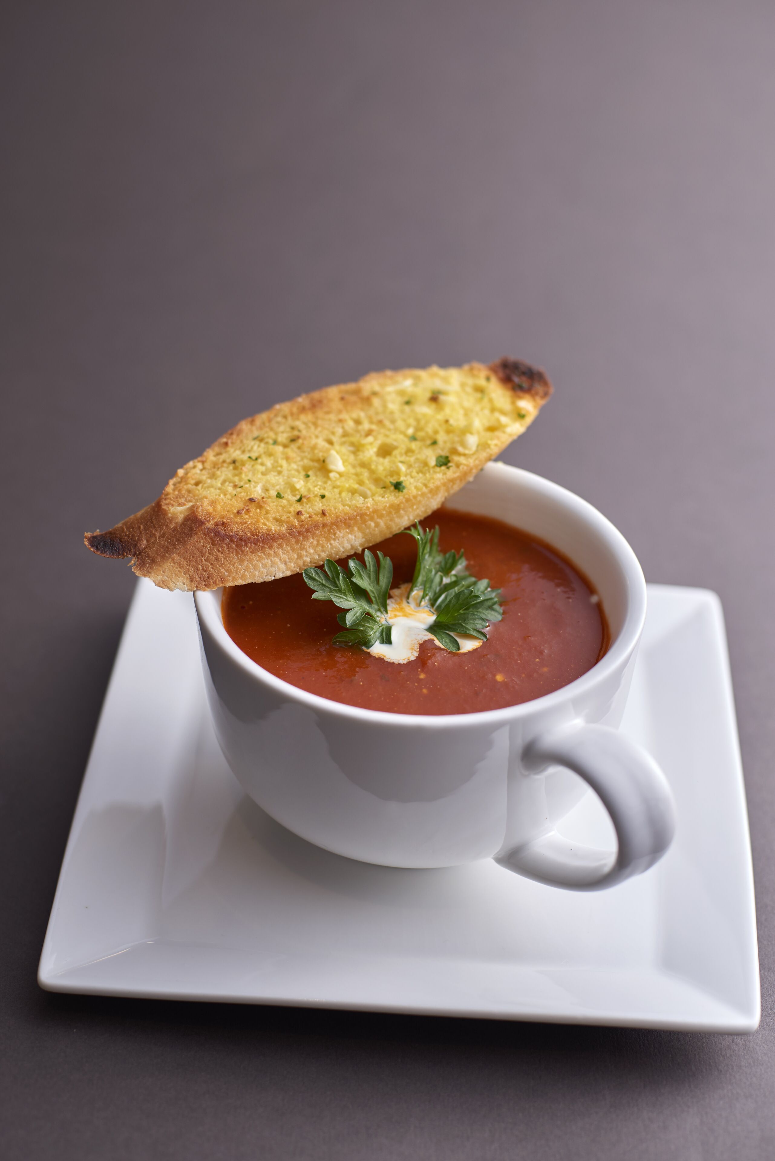A white cup of tomato soup with a piece of toasted bread on top, garnished with parsley, on a white saucer.