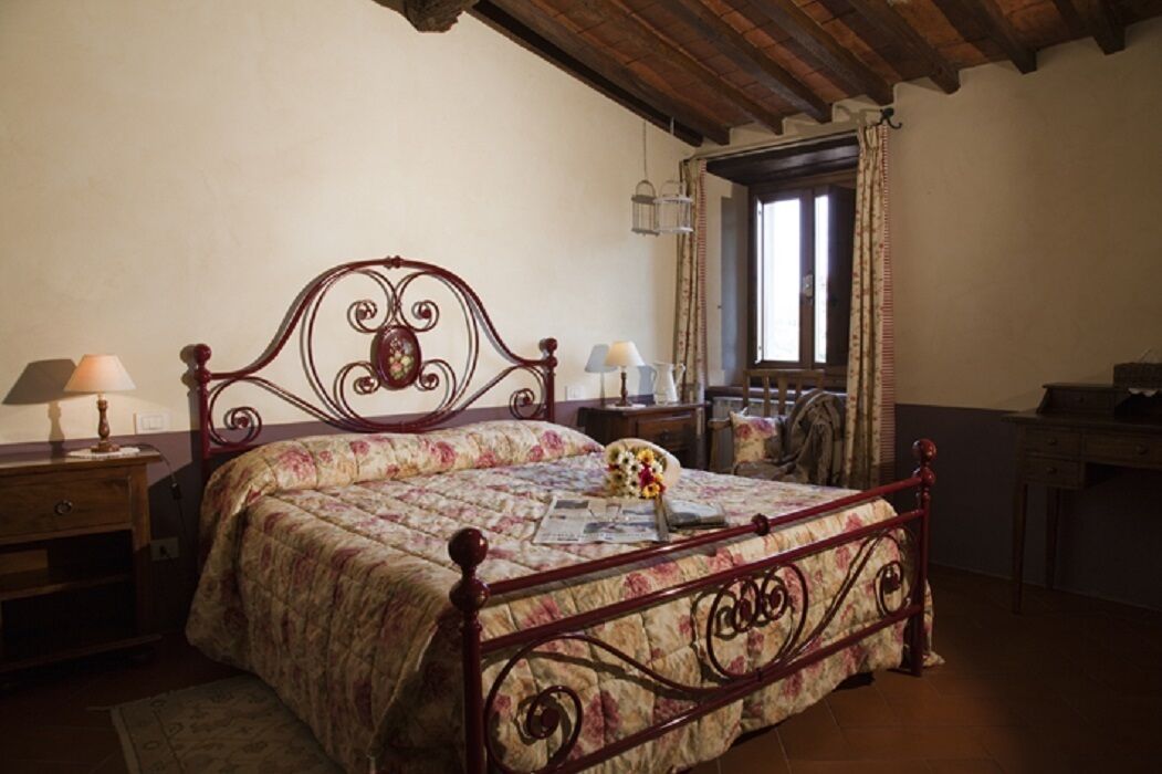 A bedroom with a wooden bed frame, floral bedding, a nightstand with a lamp, and a window with curtains.