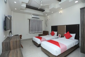 Two single beds with white bedding and red accents, a flat-screen TV, and a wooden desk with a chair.