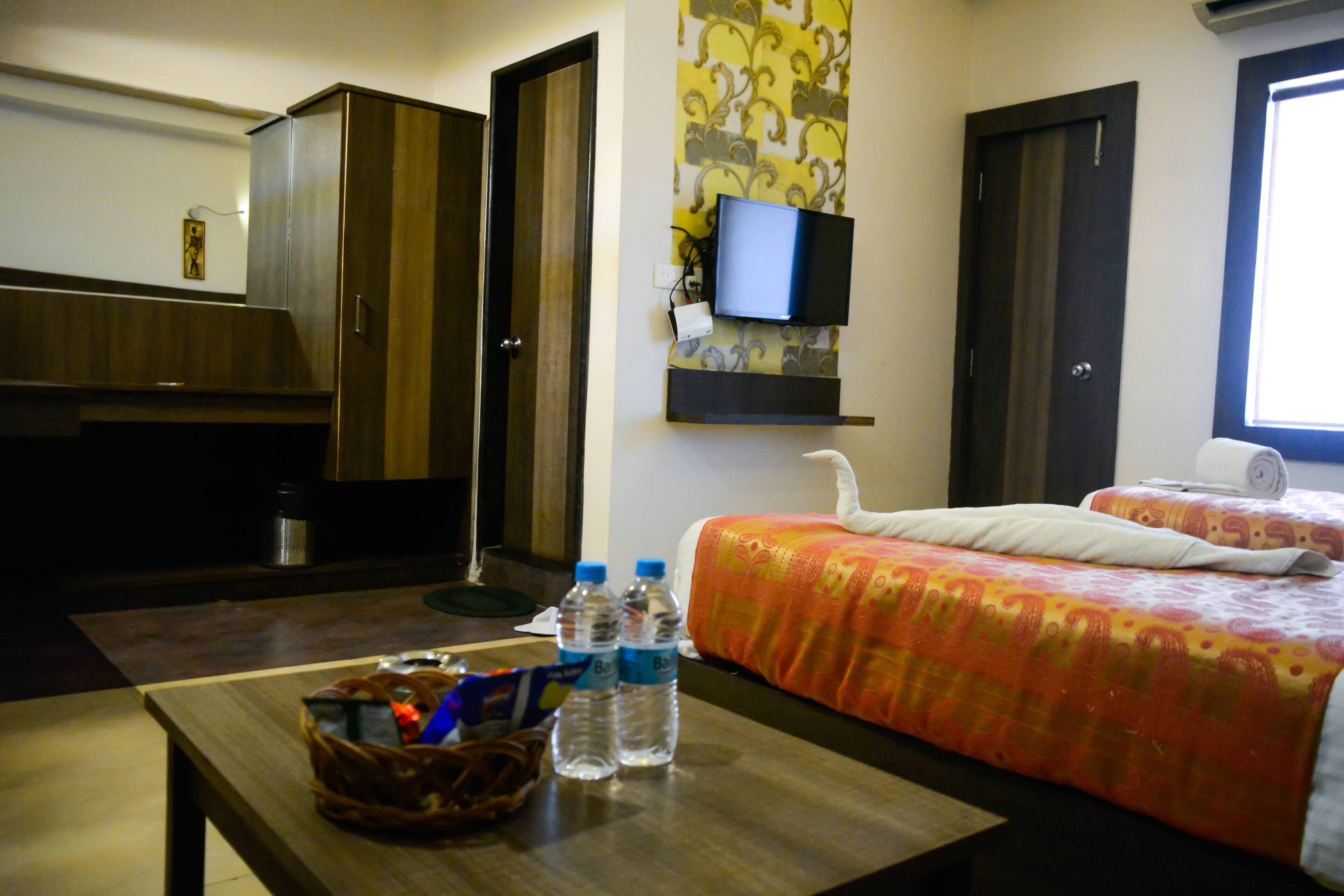 Habitación de hotel con cama, televisor montado en la pared, escritorio con una cesta y botellas de agua, y un armario.