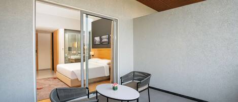 A modern hotel room with a glass door leading to a bathroom, a bed, and a small balcony with two chairs and a table.