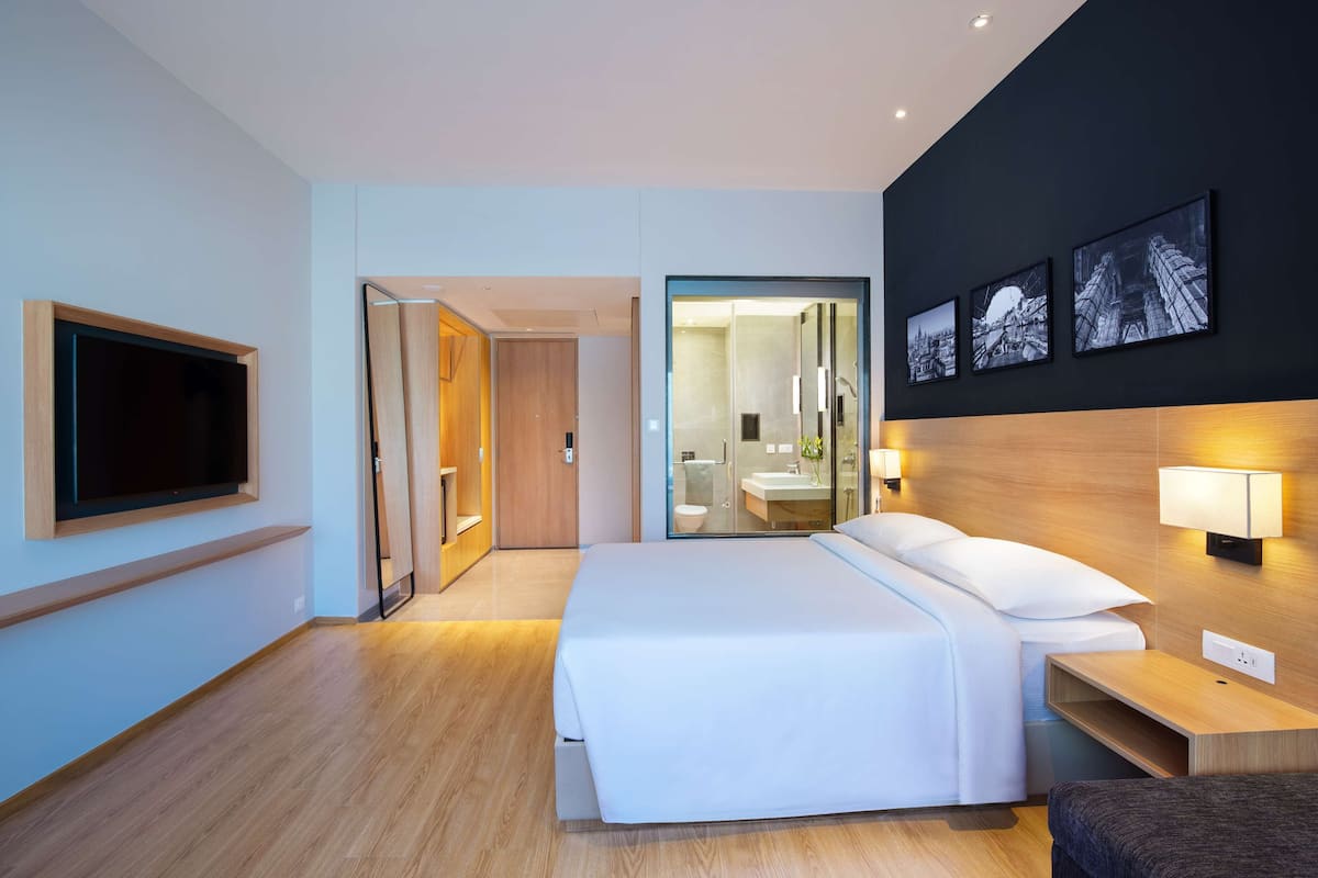 A modern hotel room with a large bed, a TV mounted on the wall, a bathroom visible through a glass door, and three framed pictures on the wall.