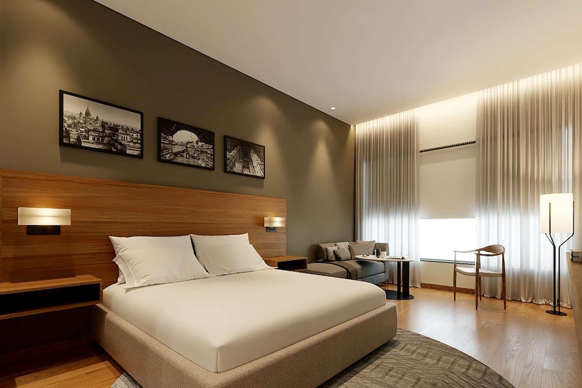 A modern hotel room with a large bed, a sofa, a small table, and a lamp. There are three framed pictures on the wall.