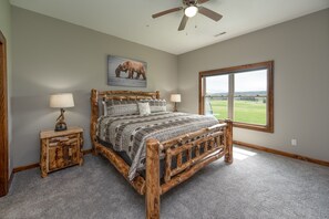 A bedroom with a wooden bed, two bedside tables, a ceiling fan, a large window, and a painting of a bear on the wall. - Luxury Dog Friendly Lodge w/ Vaulted Ceilings (Swanton)