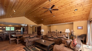 A spacious living area with a wooden ceiling, a dining table, sofas, and a kitchen in the background.
