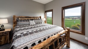 A bedroom with a large bed, wooden headboard, two bedside tables, and a view of a green landscape through a window.