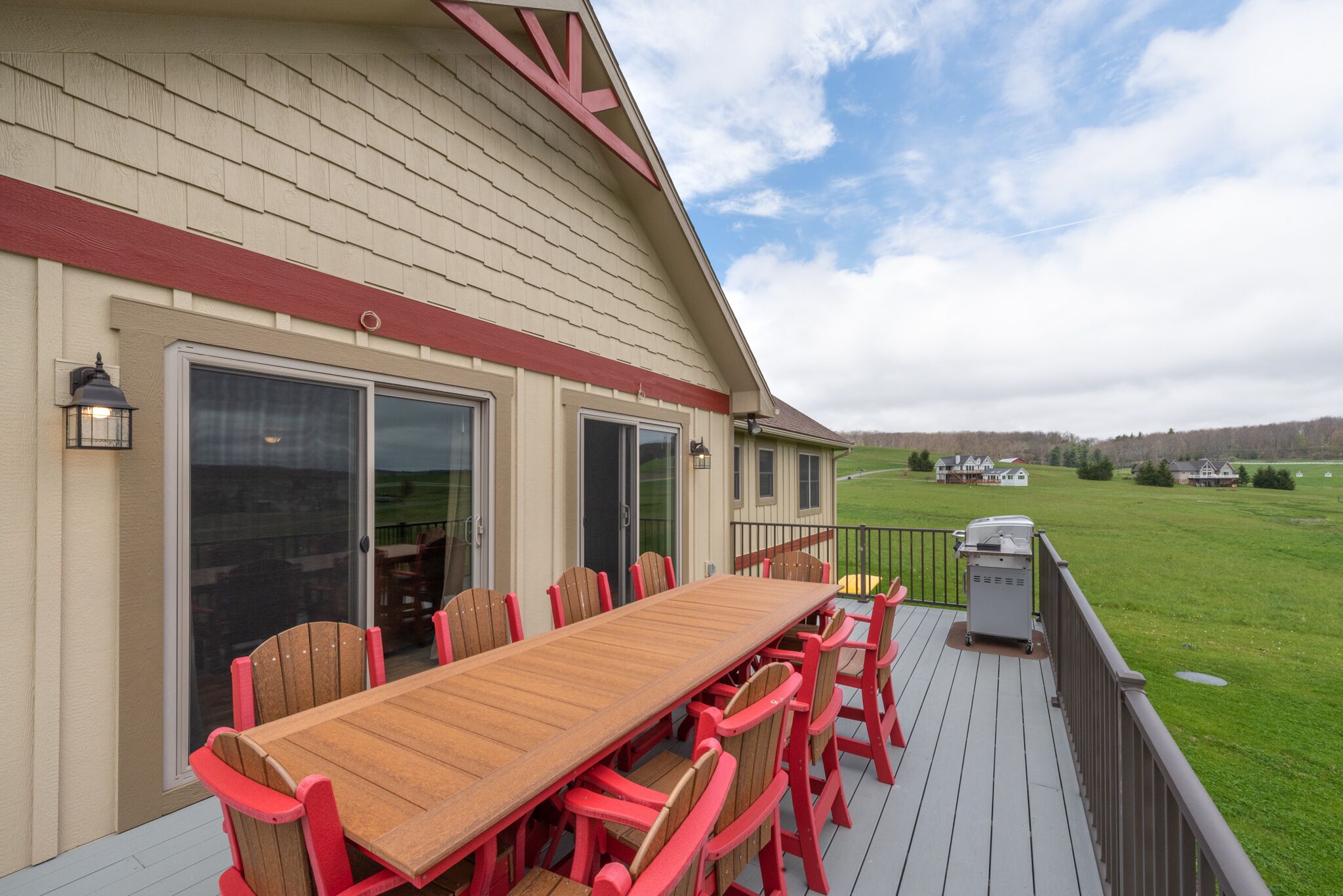 A wooden deck with a long table and red chairs, overlooking a grassy field and distant buildings.