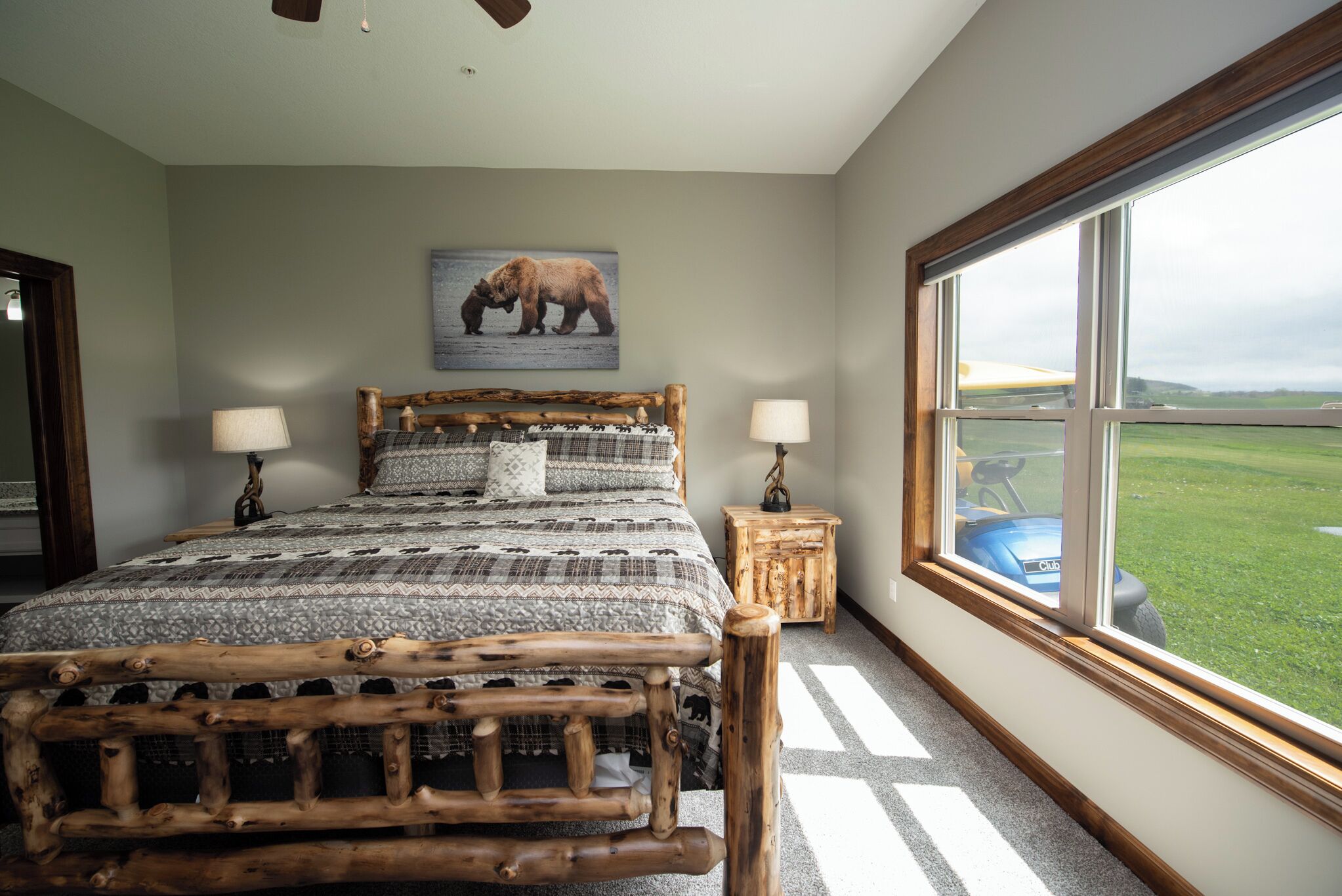 A bedroom with a wooden bed frame, a large window with a view of a green field, and a wall-mounted elephant picture.
