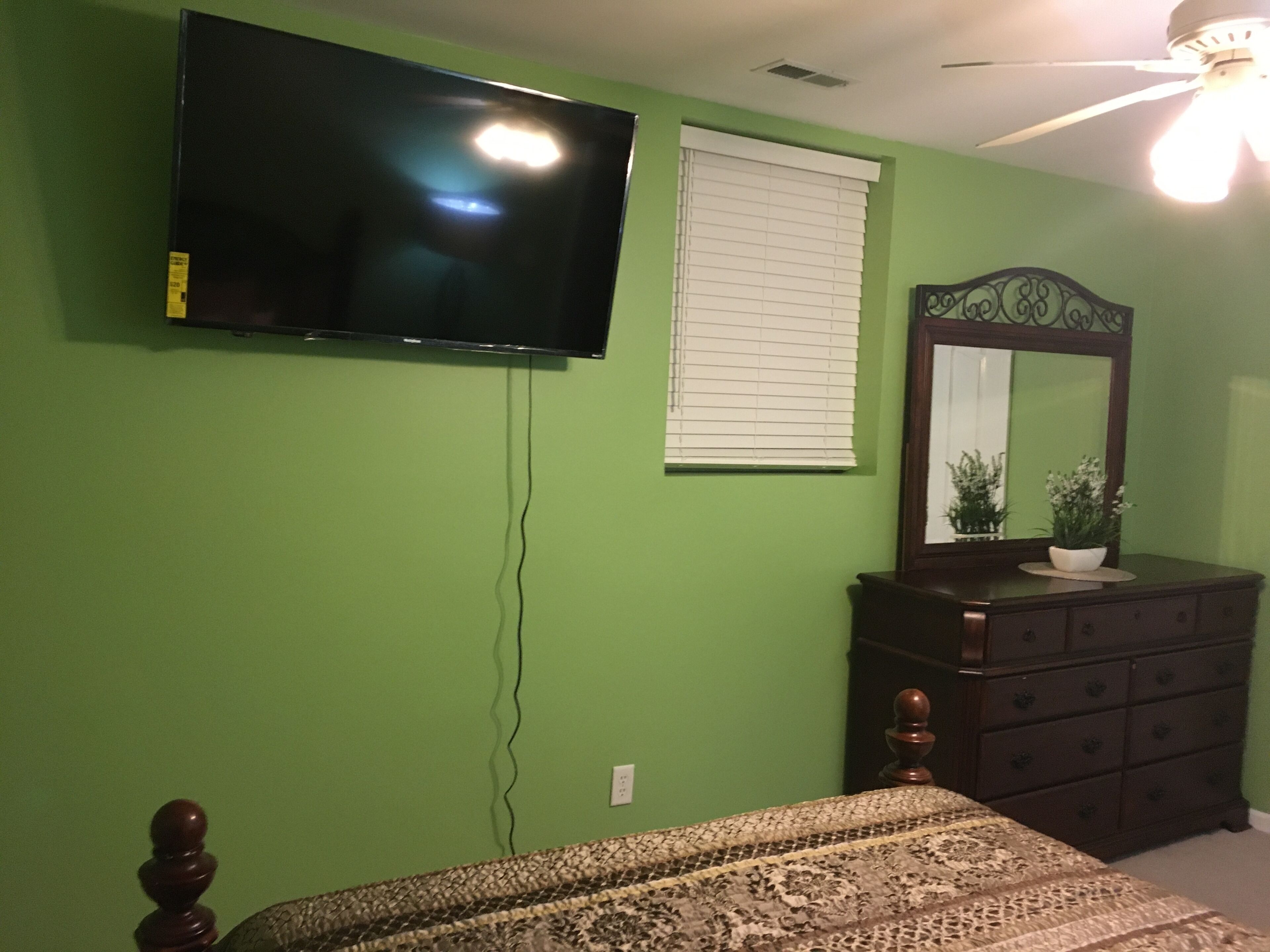 Quarto com cama, penteoudeiro, espelho e televisão presa à parede.