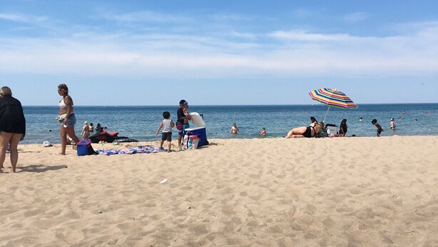 En sandstrand med folk, ett randigt parasoll och en båt i vattnet.