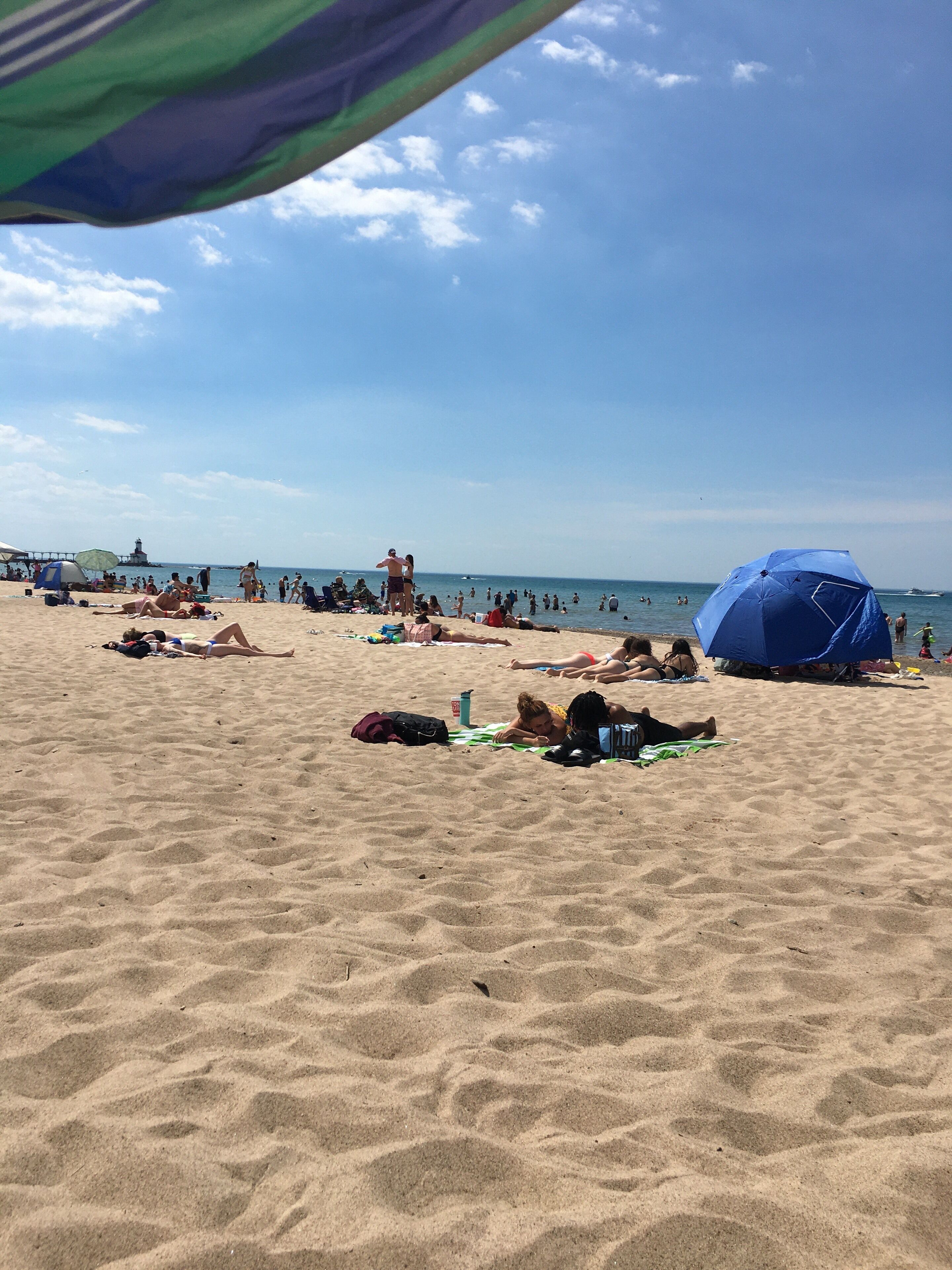 Una playa arenosa con gente tomando el sol y nadando, tiendas de campaña y un muelle al fondo.