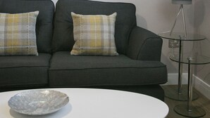 Un salon moderne avec un canapé gris, deux coussins à motifs, une table basse blanche, une table d’appoint en verre et un lampadaire.