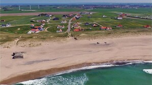 Vista aérea de un pueblo costero con casas, una playa y aerogeneradores al fondo.