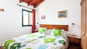 Quarto com teto de madeira, cama com colcha florida, janela com cortina, mesa de cabeceira e luminária fixada na parede.