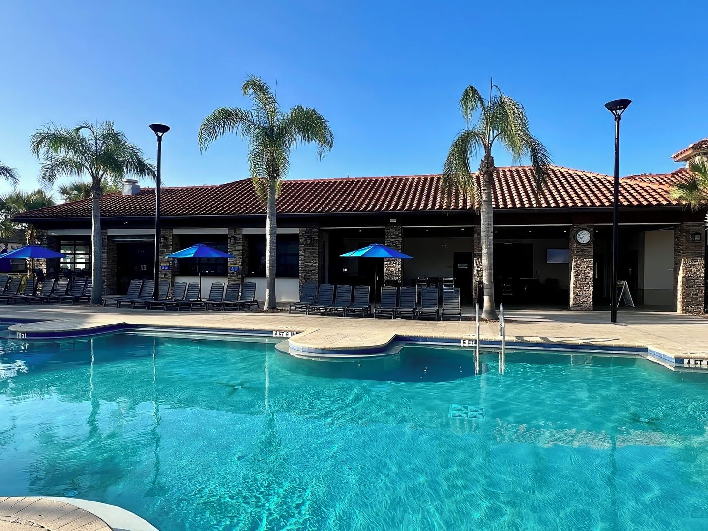 A pool area with lounge chairs and umbrellas, a building with a tiled roof, and palm trees.