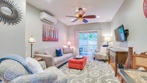 Uma sala de estar com sofá, apoio para os pés e poltrona, um ventilador de teto e uma TV fixada na parede.