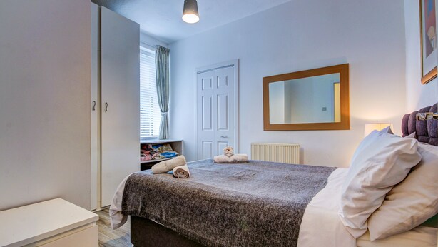 A bedroom with a bed, a bedside table, a wardrobe, a window with curtains, a door, and a mirror on the wall.