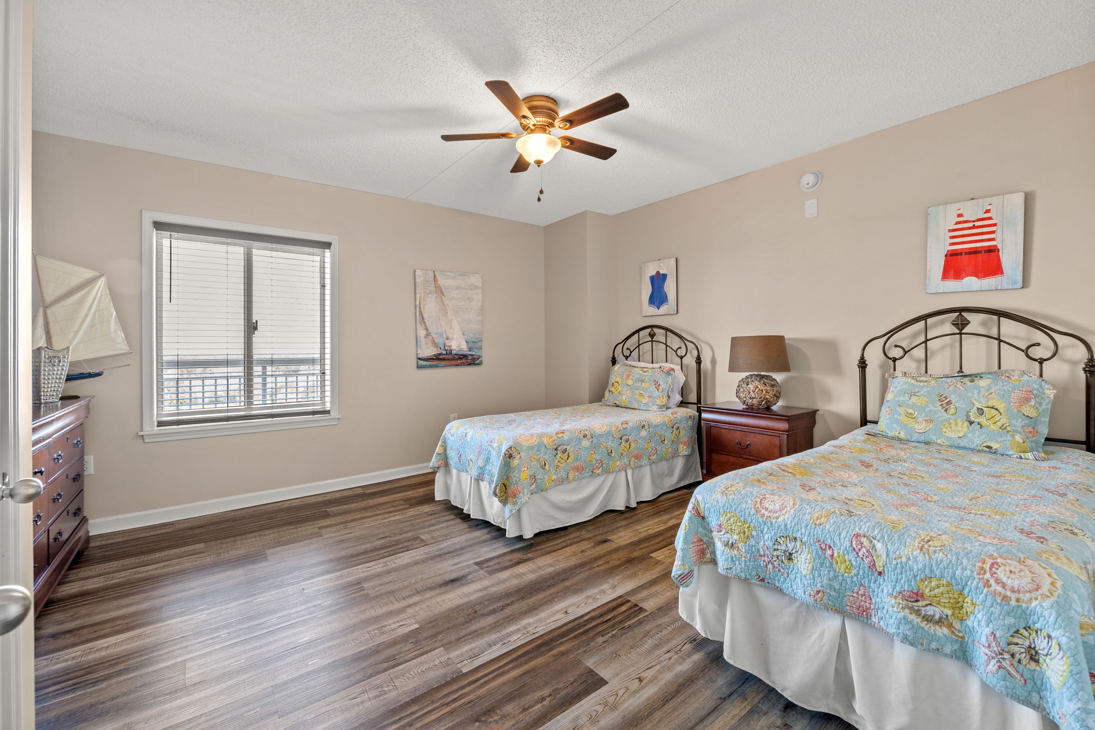 A bedroom with two beds, a ceiling fan, a window with blinds, and artwork on the walls.