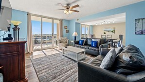 A spacious living room with a large sofa, a coffee table, and a ceiling fan. There is a sliding glass door leading to a balcony with a view of the ocean.