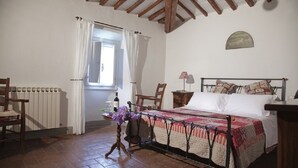 Une chambre à coucher avec un lit, une chaise, une table sur laquelle se trouvent une bouteille et des fleurs, une fenêtre avec des rideaux et un radiateur.