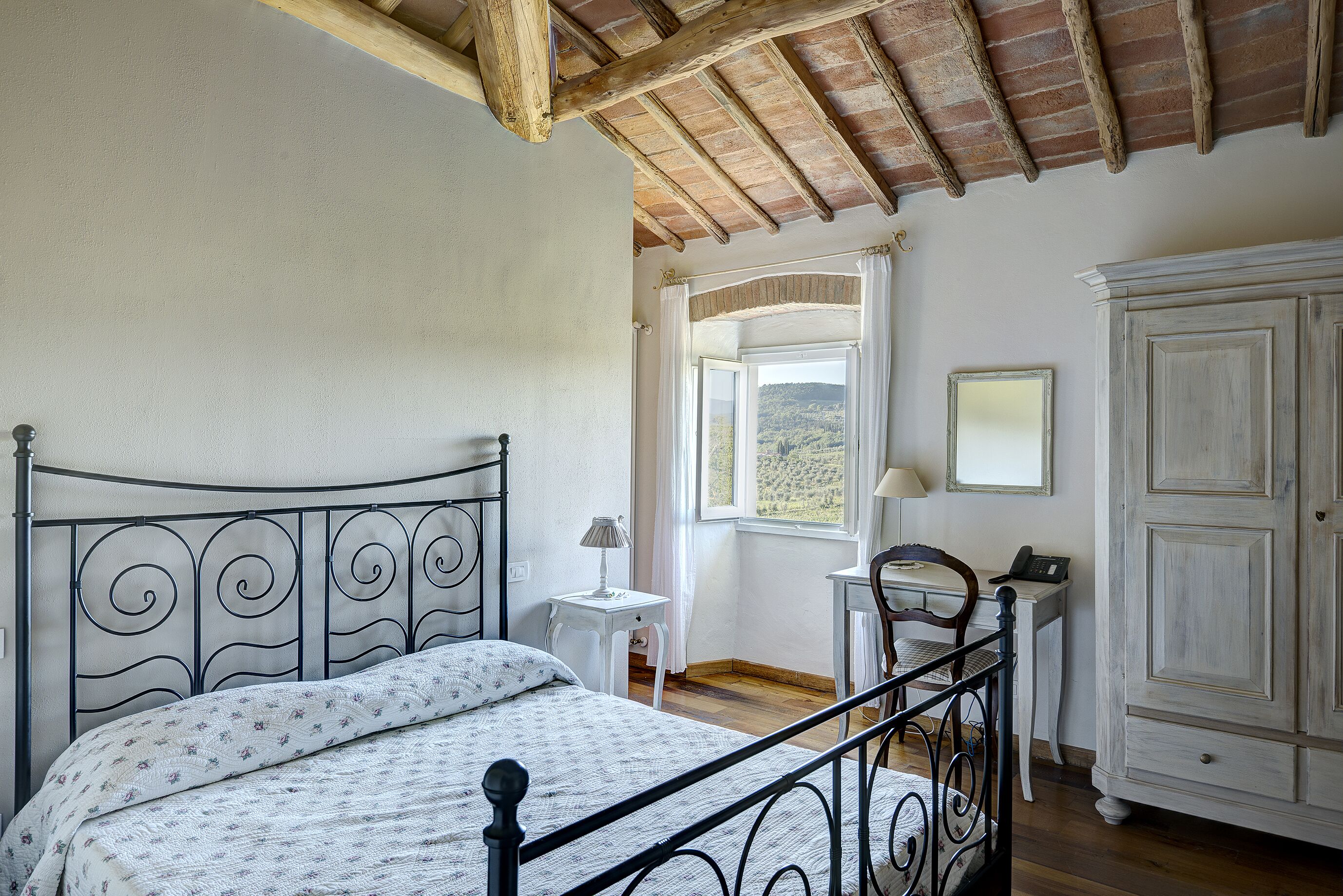 A bedroom with a metal bed frame, a white bedside table with a lamp, a wooden chair, a white wardrobe, and a window with a view of the countryside.