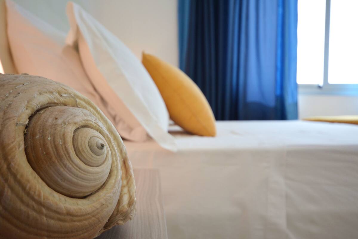 A bed with white bedding and a yellow pillow, a wooden headboard, and a window with blue curtains.