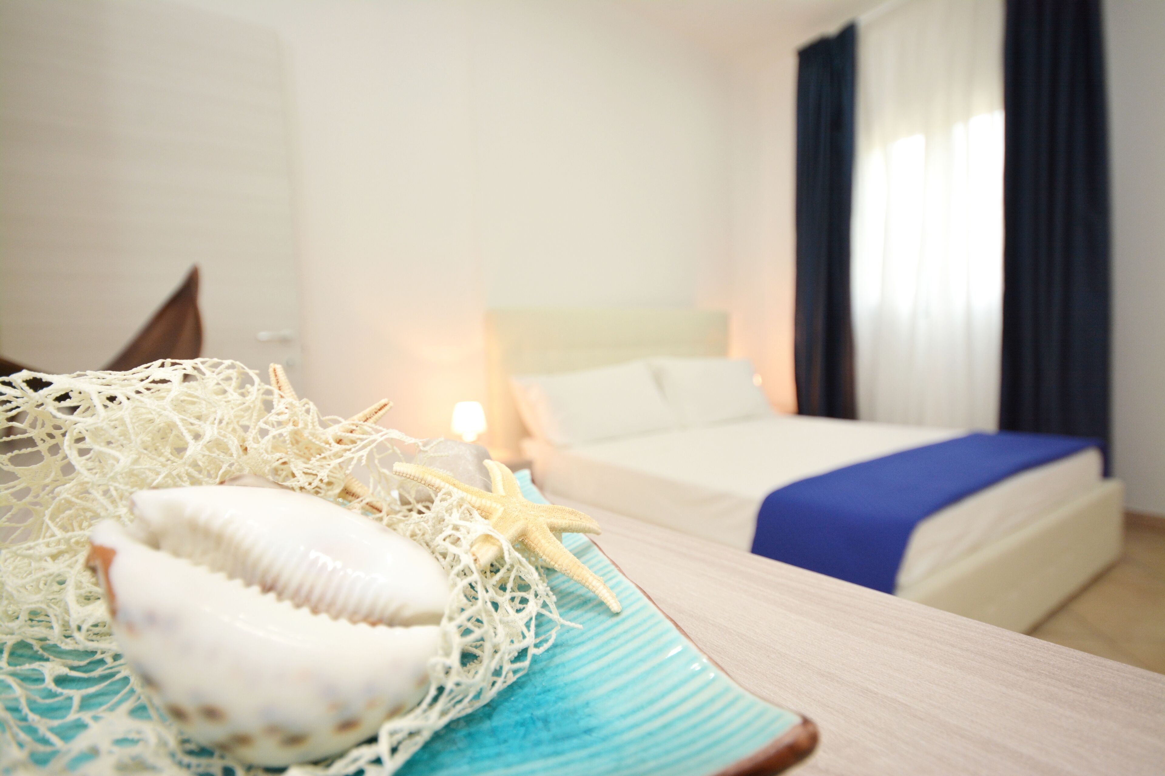 A hotel room with a bed, a bedside table with a decorative shell and starfish, and a window with curtains.