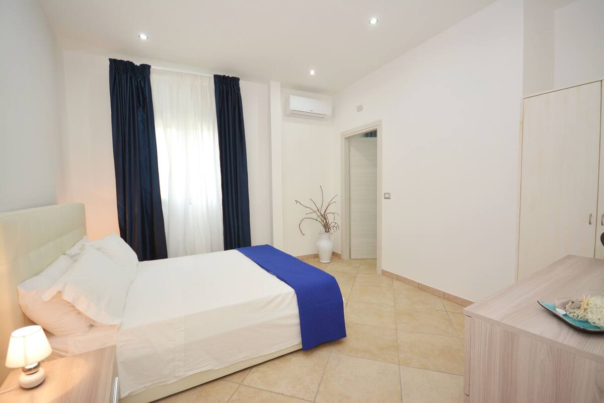 A hotel room with a bed, bedside table, lamp, and a view of the hallway.