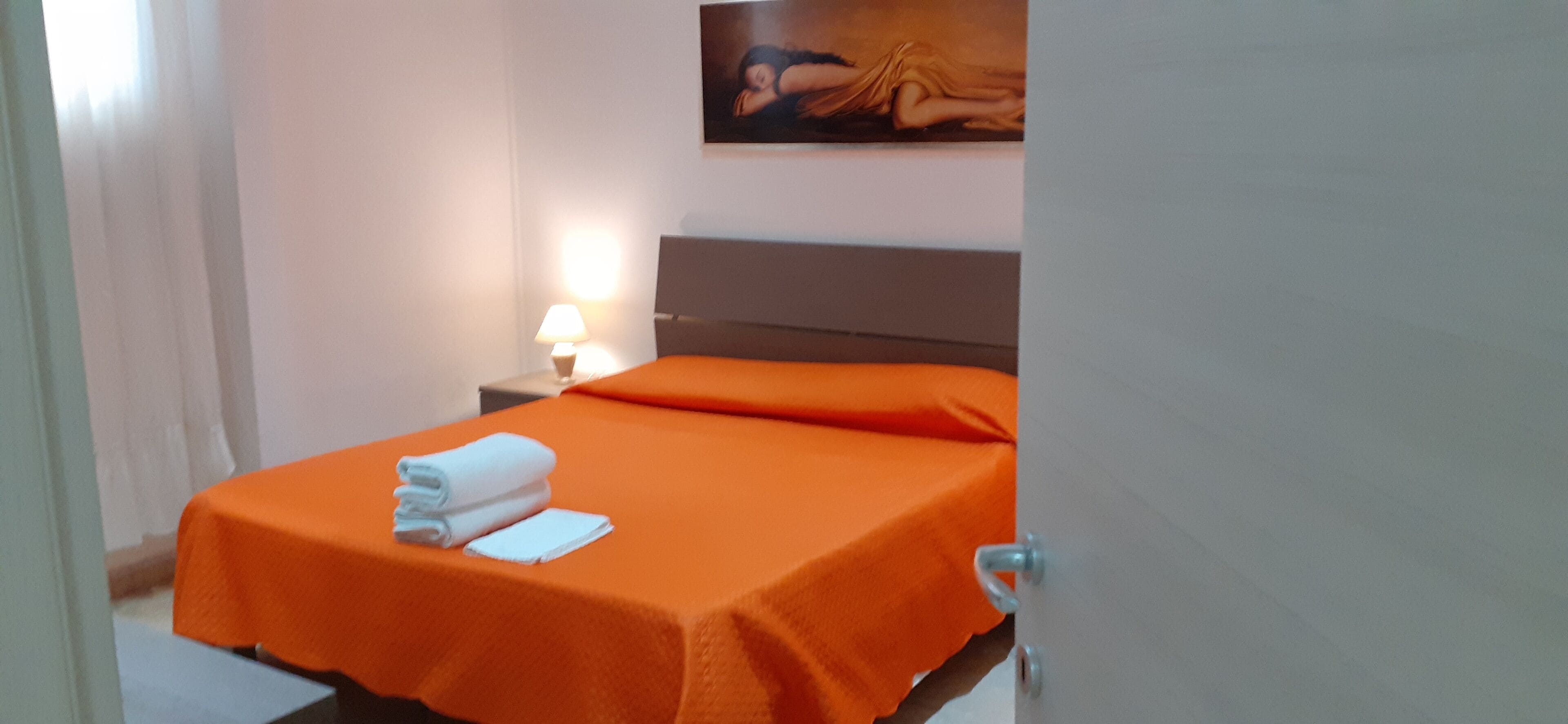 A hotel room with a bed covered in orange bedding, a nightstand with a lamp, and a painting on the wall.
