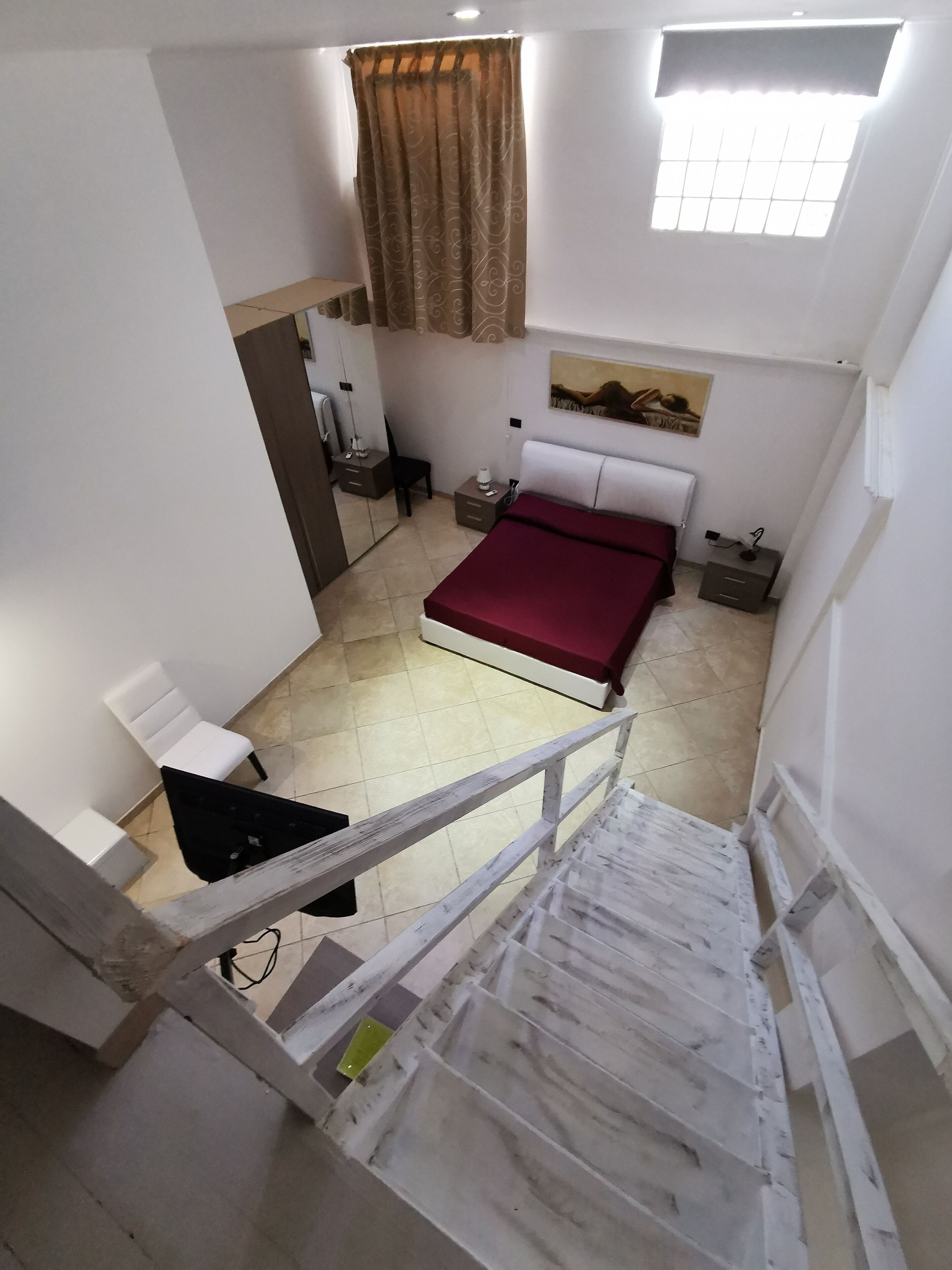 Un dormitorio con cama, mesita de noche, silla, ventana con cortinas y un cuadro en la pared.